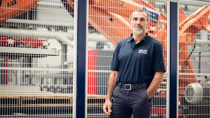 Markus Schopp, wearing a dark blue polo shirt with the BIKAR METALS logo, stands in a production hall in front of a large orange robotic arm that is moving aluminum cut pieces.