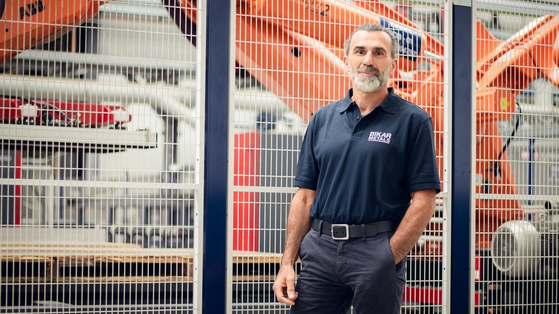 Markus Schopp im dunkelblauem Poloshirt mit BIKAR METALS Logo steht in einer Produktionshalle vor einem großen, orangefarbenen Roboterarm, der Aluminiumzuschnitte bewegt.