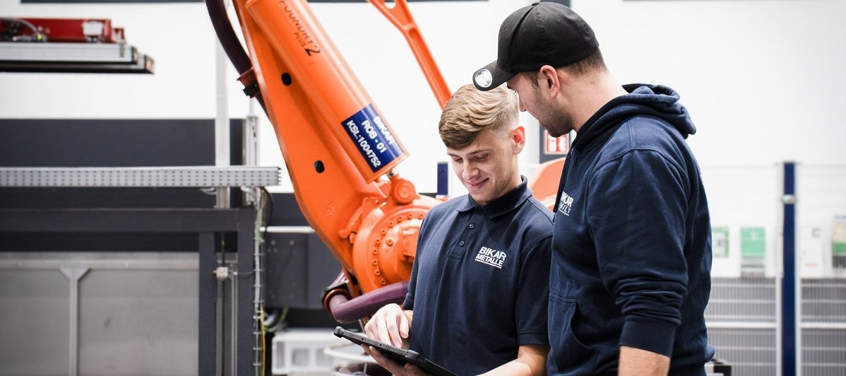 Ein Ausbilder erklärt einem Auszubildenden an einem Tablet die Arbeit mit einem orangefarbenen Industrieroboter bei BIKAR METALS in Bad Berleburg.
